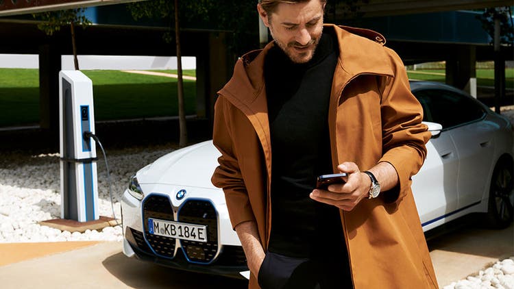 BMW Service Inclusive čovek gleda u svoj pametni telefon ispred BMW-a i4 na stanici za punjenje